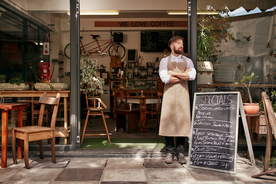 Portrait of a young man standing in front of his coffee shop. Young man with beard wearing an apron standing with her arms crossed and looking away.